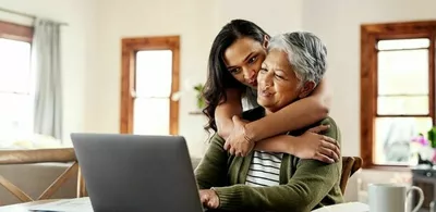 En eldre dame sitter med en laptop og en yngre dame klemmer henne bakfra