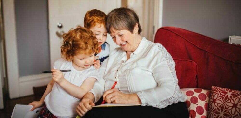 En eldre dame sitter med to barn i en sofa og skriver på et ark