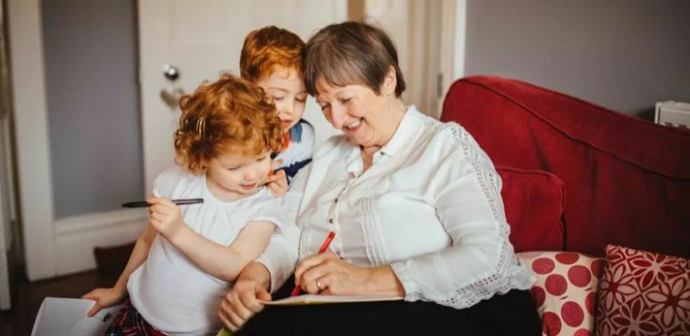 En eldre dame sitter med to barn i en sofa og skriver på et ark